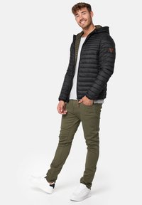 Black puffer jacket with a hood and olive green lining, paired with olive green pants and white sneakers, standing against a white background.