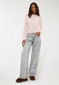 Maglione rosa chiaro con maniche lunghe, abbinato a jeans a gamba larga azzurri a vita alta e scarpe marroni scure, esposto su uno sfondo bianco.