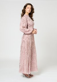 Langes pinkes Kleid mit hohem Kragen, transparenten langen Ärmeln und verzierten Pailletten-Details. Es bietet ein fließendes Design und einen subtilen Glanz. Silberne High Heels.
