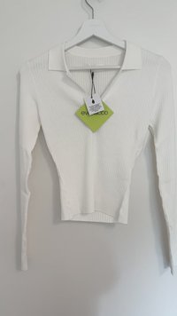 Bijela majica dugih rukava s rebrastom teksturom, V-izrezom i ravnim ovratnikom. Uključuje zeleni tag s imenom brenda "ever&odd".