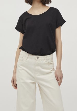 Nainen, jolla on yllään löysä musta lyhythihainen paita, joka on sujautettu korkeavyötäröisiin, leveälahkeisiin, luonnonvalkoisiin housuihin, seisoo yksiväristä taustaa vasten.