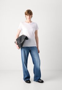 T-shirt bianco a maniche corte con un piccolo logo, abbinata a dei jeans blu larghi e scarpe di pelle nere, mentre tiene una borsa di pelle nera.