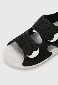 Czarna klapka na płaskiej podeszwie z fakturowaną górą, wyposażona w dwa regulowane paski i białe akcenty, na białej miękkiej podeszwie z żłobionym wzorem.