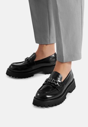 Schwarze Loafer aus Lackleder mit dicksohliger schwarzer Sohle, verziert mit dekorativem Metallbeschlag am Obermaterial und einer glatten, glänzenden Oberfläche.