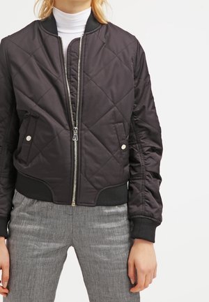 Kobieta w czarnej pikowanej bomberce zapinanej na zamek, na białym golfie i szarych spodniach na jednolitym tle.
