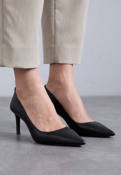 Escarpins en cuir noir à bout pointu, avec un talon fin et une texture lisse, portés avec un pantalon léger et bien ajusté.