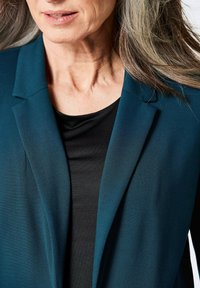 Teal blazer met een gladde textuur, met geknipte revers, gedragen boven een zwarte top, wat een gelaagd effect en een getailleerde pasvorm accentueert.