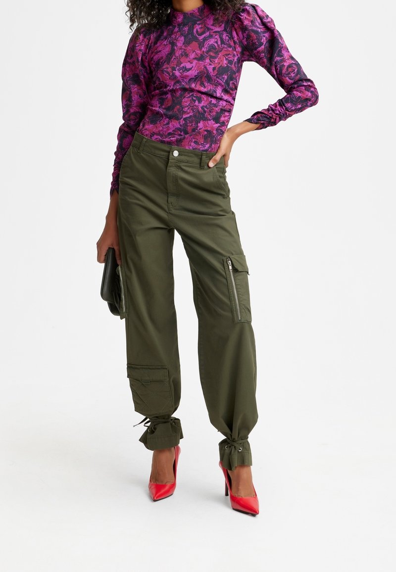 Femme portant un haut à manches longues violet à motifs, un pantalon cargo vert olive avec des poignets noués, des talons hauts rouges et tenant une petite pochette.