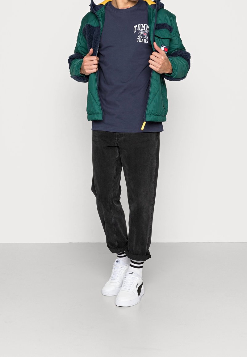 Zelená prošívaná bunda s námořnickým fleecovým podšívkou, námořnické tričko s nápisem, černé zúžené kalhoty a bílé tenisky s černými detaily.