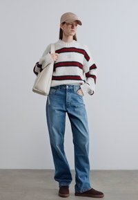 Λευκό και κόκκινο ριγέ πουλόβερ, μπλε τζιν, μπεζ καπέλο και κρεμ τσάντα ώμου. Καφέ παπούτσια. Casual εμφάνιση σε ουδέτερο φόντο.