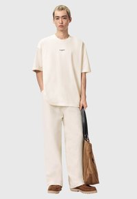 Beige bomulls-T-shirt med korta ärmar, med en liten svart logotyp. Kompletterad med matchande löst sittande byxor och bruna slip-in-skor.