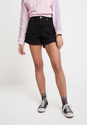Person trägt schwarze hochgekrempelte Jeans-Shorts, einen pinken Hoodie mit violetten Streifen an den Ärmeln, Socken mit Leopardenmuster und schwarze Sneakers mit weißen Schnürsenkeln.