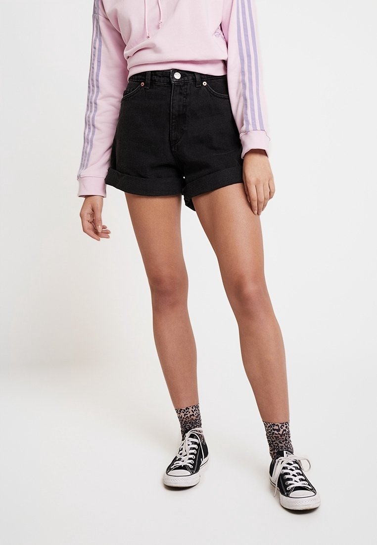 Personne portant un short en jean noir retroussé, un sweat à capuche rose avec des rayures violettes sur les manches, des chaussettes imprimé léopard et des baskets noires avec des lacets blancs.