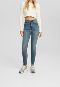 Jeans skinny de tiro alto en azul claro con bolsillos frontales, combinados con un suéter ajustado de canalé blanco y zapatillas grises.