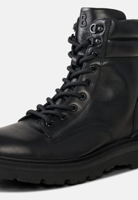 Černé kožené kotníkové boty s kulatou špičkou, šněrováním v přední části a robustní podrážkou. Obsahují prošívání a logo na horní straně.