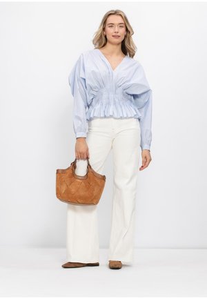 Vrouw draagt een lichtblauwe blouse met plooien, witte broek met wijde pijpen, bruine schoenen, en houdt een bruine leren handtas vast tegen een witte achtergrond.