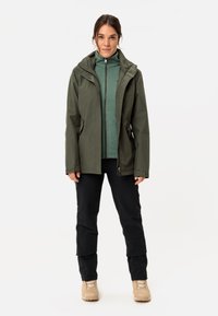 Giacca verde oliva resistente all'acqua con zip frontale, cappuccio e tasche laterali, indossata sopra una felpa con zip verde chiaro e pantaloni neri.