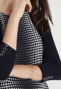 Nainen, joka pukeutuu mustavalkoiseen polkadot-kuvioituun mekkoon, mustaan pitkähihainen neuletakkiin ja hopeiseen geometriseen rannekoruun.