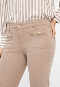 Beige, eng anliegende Hose mit glatter Textur, goldenen Knopfakzenten und zwei hinteren Taschenklappen. Gestylt mit einer hell gestreiften Bluse.