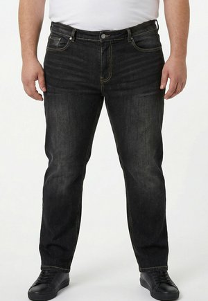 Mann trägt schwarze ausgewaschene Jeans mit gelber Naht, schwarze Turnschuhe und ein weißes Hemd, steht vor einem weißen Hintergrund.