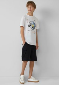 Weißes Baumwoll-T-Shirt mit Wellenmotiv in Blau und Gelb, kombiniert mit schwarzen Cargo-Shorts. Weiße Sneakers mit sandfarbenen Sohlen und Akzenten.