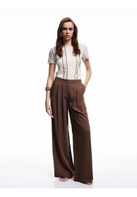 Jasny, fakturowany top z krótkimi rękawami i pionowymi paskami, w zestawieniu z szerokimi, brązowymi spodniami. Uzupełniony drewnianą bransoletką.