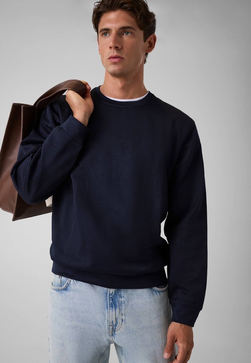 Homme portant un pull texturé bleu marine foncé, un jean bleu clair, tenant un grand sac en cuir marron sur son épaule, regardant sur le côté.