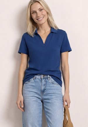 Glimlachende vrouw met een blauw poloshirt met korte mouwen en lichtblauwe spijkerbroek, houdt een beige handtas vast tegen een effen lichte achtergrond.