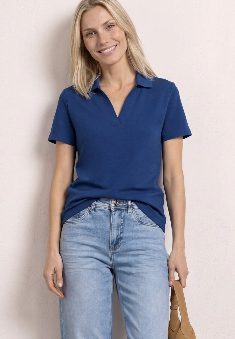 Donna sorridente che indossa una polo blu a maniche corte e jeans azzurri, che tiene una borsa beige davanti a uno sfondo chiaro uniforme.