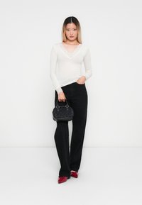 Femme portant un haut blanc avec des dentelles aux manches, un pantalon noir, des chaussures à talons rouges, tenant un petit sac à main noir, sur fond blanc.