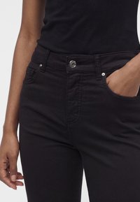 Femme portant un pantalon noir et un haut noir sans manches, une main dans une poche et l'autre détendue le long du corps.
