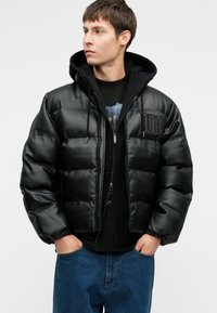 Jovem vestido com um casaco acolchoado de couro preto por cima de um hoodie preto e jeans azuis, com as mãos nos bolsos, olhando para o lado.