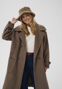 Rjav volnen plašč z dvojim zapenjanjem in velikimi reverji ter sprednjimi žepi, v kombinaciji s svetlobržem teksturiranem klobuku in belim puloverjem.