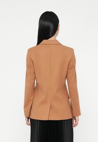 Blazer marrone su misura realizzato in tessuto morbido, con colletto a reverse e una fessura singola sul retro. Chi lo indossa è di profilo, con capelli scuri.