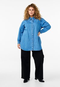 Camicia button-up in denim blu chiaro con maniche lunghe, caratterizzata da un motivo testurizzato e un orlo curvo. Abbinata a pantaloni neri larghi e stivali alla caviglia.