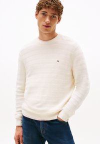 Cremefarbener Pullover mit strukturierten horizontalen Streifen, geripptem Kragen und Bündchen, kleinem gestickten Logo auf der Brust, kombiniert mit blauen Jeans.