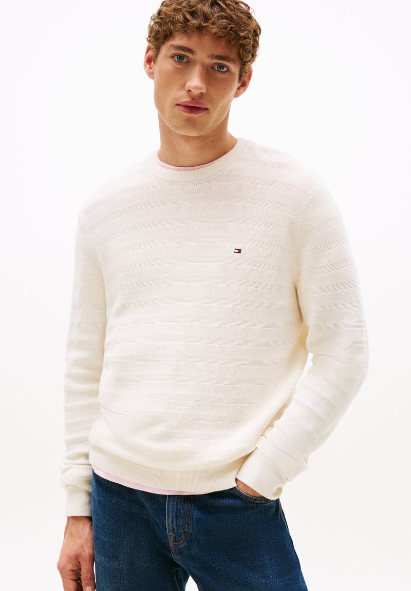 Cremefarbener Pullover mit strukturierten horizontalen Streifen, geripptem Kragen und Bündchen, kleinem gestickten Logo auf der Brust, kombiniert mit blauen Jeans.