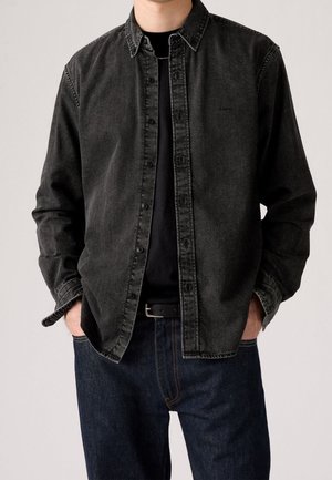 Man wearing a black denim button-up shirt over a black t-shirt with dark blue jeans and hands in pockets, shown from shoulders to hips.