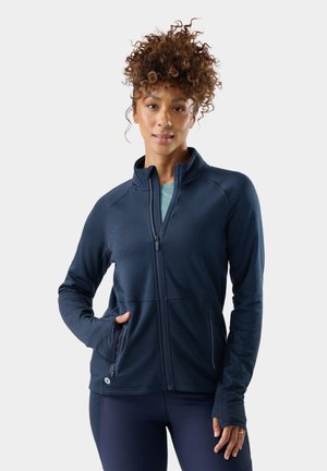 Mujer con cabello rizado que lleva una chaqueta azul marino con cremallera y leggings, de pie con una mano en el bolsillo de la chaqueta.