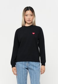 Black long-sleeve shirt with ribbed cuffs featuring a small red heart logo on the chest, paired with light blue vertically striped jeans.