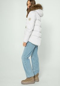 Bílá péřová bunda s kapucí z umělé kožešiny, s zlatým emblémem na rukávu, v kombinaci se světle modrými džíny a béžovými botami.
