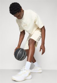 Lys beige atletisk outfit, der inkluderer en kortærmet top og shorts. Hvide sneakers med guld accenter. Holder en sort basketball.