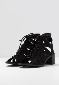 Czarne zamszowe sandały na obcasie z odkrytymi palcami charakteryzują się wyciętymi wzorami, sznurowanym przodem oraz teksturowanym blokowym obcasem dla zwiększonej stabilności.