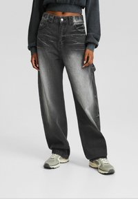 Person som bär urtvättade svarta vida jeans, kort mörk sweatshirt och ljusgrå sneakers mot en enfärgad grå bakgrund.