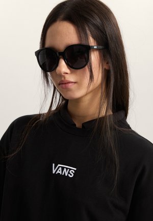Gafas de sol negras de gran tamaño con montura redonda, complementadas por una camisa negra con el logo blanco de Vans. El cabello texturizado cae alrededor del rostro.