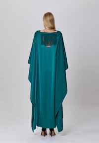 Robe cape en satin teal, au design fluide, avec un dos découvert orné de franges et des fentes hautes sur les côtés, associée à des stilettos noirs.
