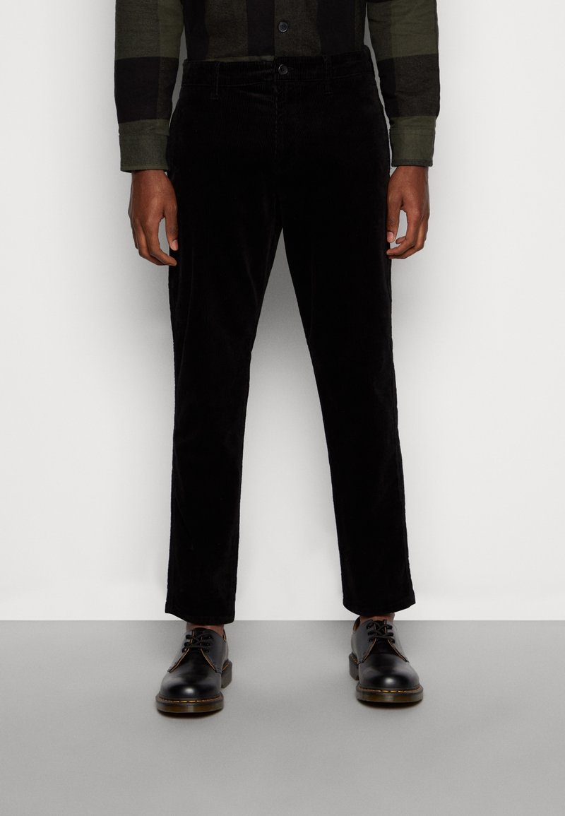 Homme portant un pantalon en velours côtelé noir, des chaussures en cuir noires à lacets et une chemise à carreaux vert foncé et noir à manches longues, debout sur un sol gris.
