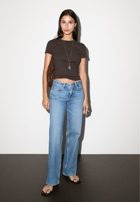 Hnědý přiléhavý tričko, modré rozšířené jeansy s vysokým pasem. Model drží texturovanou hnědou tašku, má na sobě černé žabky. Jednoduchý náhrdelník.