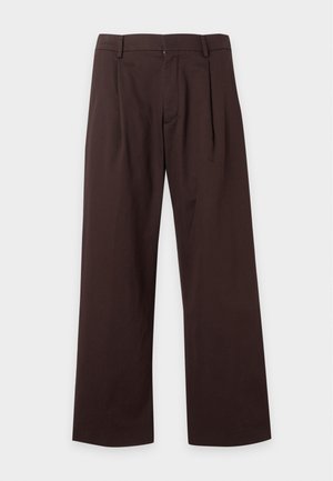 Jeune homme portant un pull marron clair par-dessus une chemise blanche, pantalon large marron foncé, baskets blanches, debout sur un fond blanc uni.