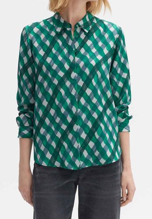 Kobieta ubrana w zielono-białą koszulę w kratę z podwiniętymi rękawami i ciemnoszare spodnie, stojąca na jednolitym tle.